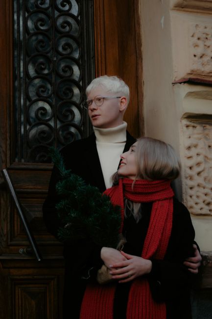 A happy couple stands in front of a decorative door holding a small holiday tree.