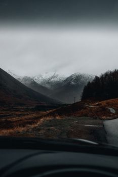 A foggy landscape with snowy mountains seen from a vehicle window on a winter day.