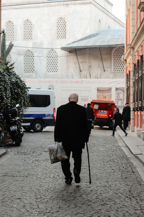 Free A senior adult man walks with a cane on a cobblestone street in a town, carrying a bag. Stock Photo