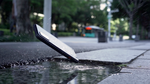 Free A smartphone splashes into a city puddle, capturing motion and water interaction. Stock Photo