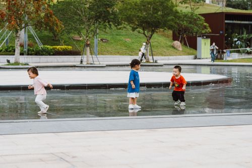 Free Three children playing joyfully on a wet park pavement, surrounded by greenery and trees. Stock Photo