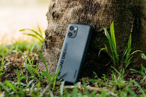 Free Close-up of a smartphone resting against a tree trunk amidst grass, showcasing vibrant outdoor life. Stock Photo