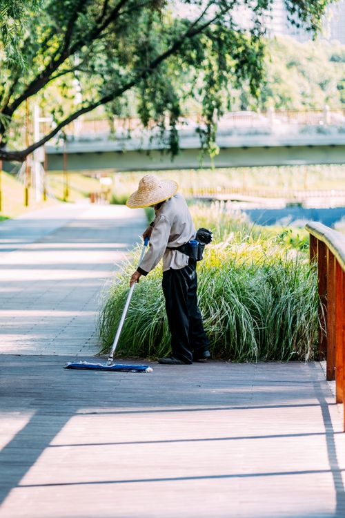 Free A park worker wearing a straw hat sweeps a wooden pathway surrounded by greenery. Stock Photo