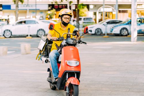 Free A delivery courier rides a red scooter on a city street, focused on navigating urban traffic. Stock Photo