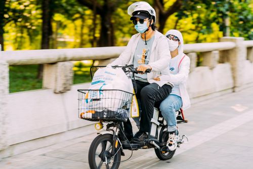 Free A couple in face masks riding a scooter with shopping on a sunny street. Stock Photo