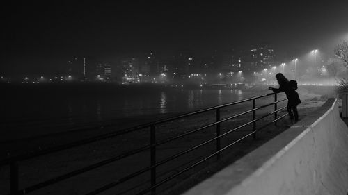 Free A solitary silhouette of a woman leaning on a railing by a foggy city waterfront at night. Stock Photo