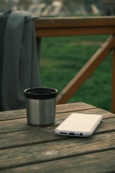 A smartphone and metal cup rest on a rustic wooden table outdoors, creating a serene and modern atmosphere.