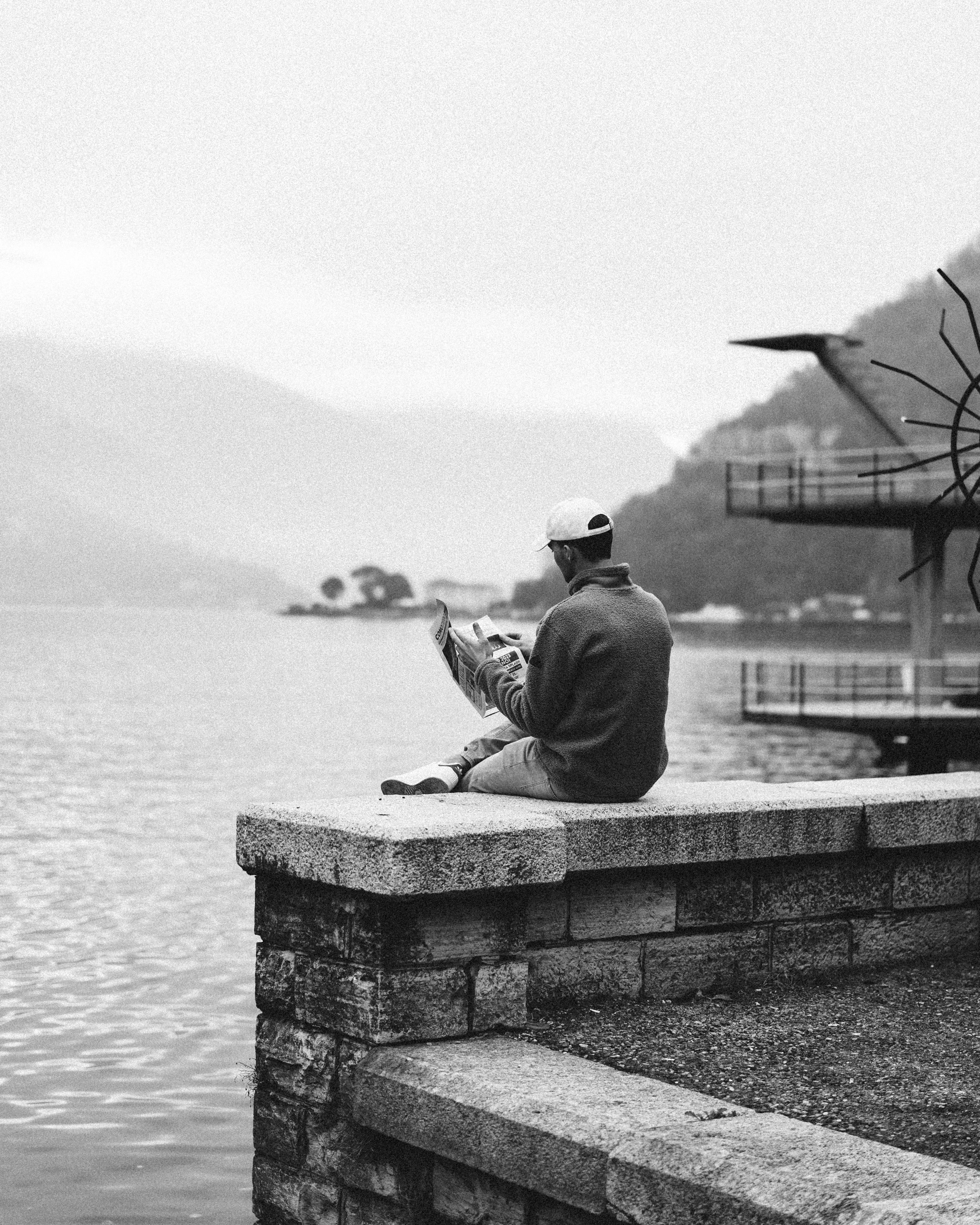 A man reading a newspaper while sitting by Lake Como, Italy in a tranquil black and white setting.