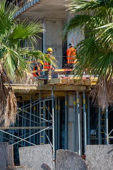 Construction workers in safety vests on scaffolding with palm trees in the foreground.