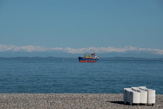 Cargo ship sailing on calm sea against a backdrop of majestic mountains under a clear blue sky.
