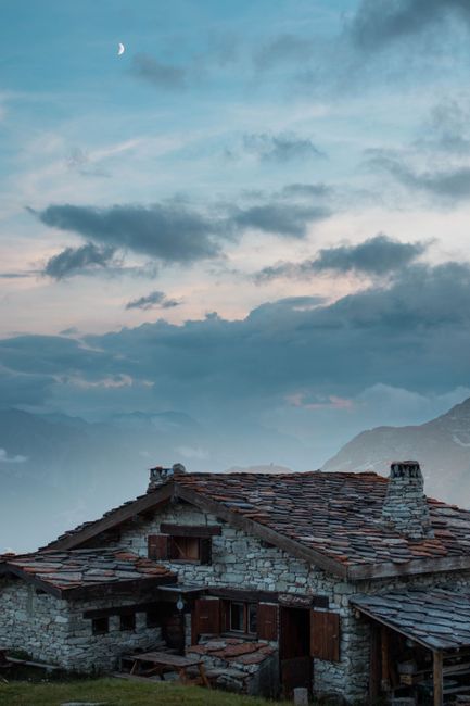 Charming stone house with chimneys under a serene dusk sky in a mountainous area, featuring clouds and a crescent moon.