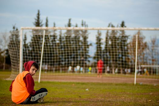 A boy sits cross-legged on a football field, wearing a hoodie and an orange vest.
