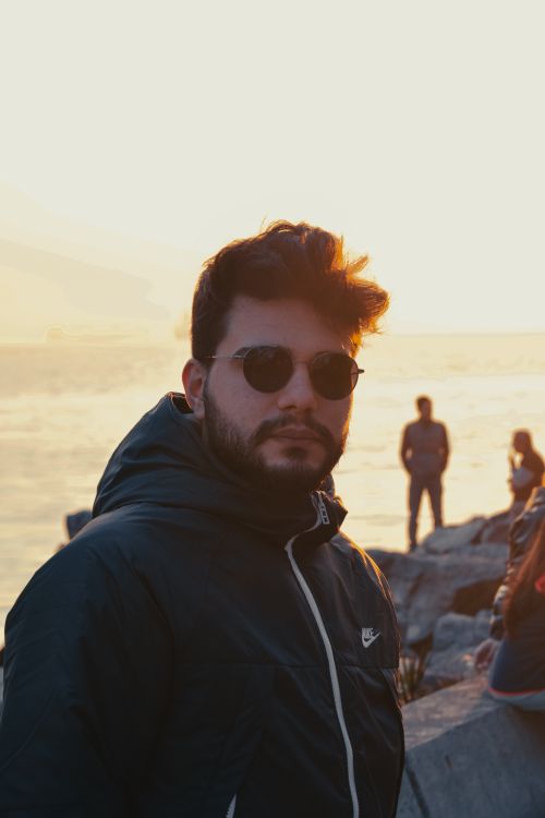 Free Man wearing sunglasses in front of a scenic sunset at the seaside, with people in the background. Stock Photo
