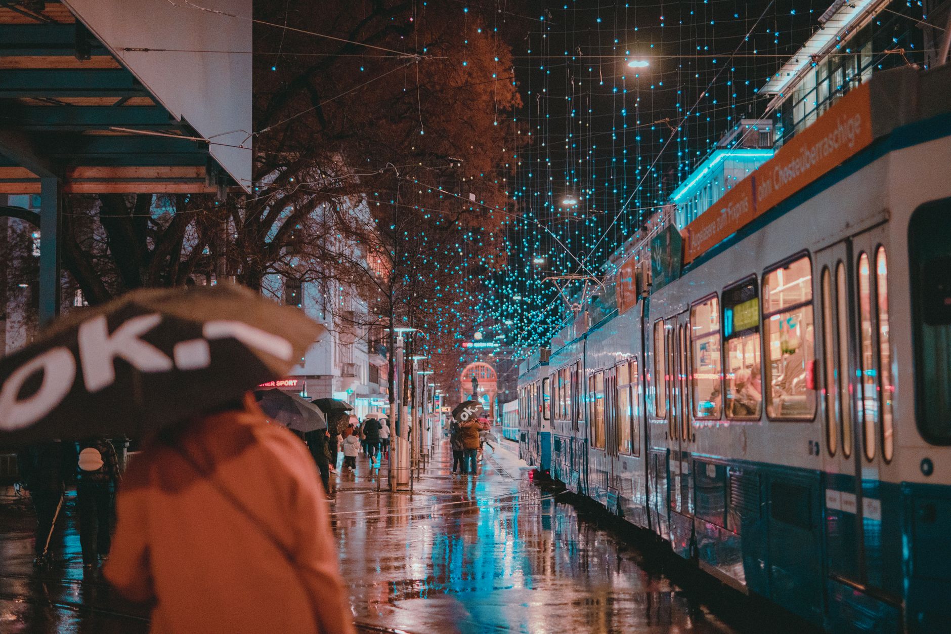Moody rainy night in Zürich with trams and people with umbrellas under illuminated streetlights.