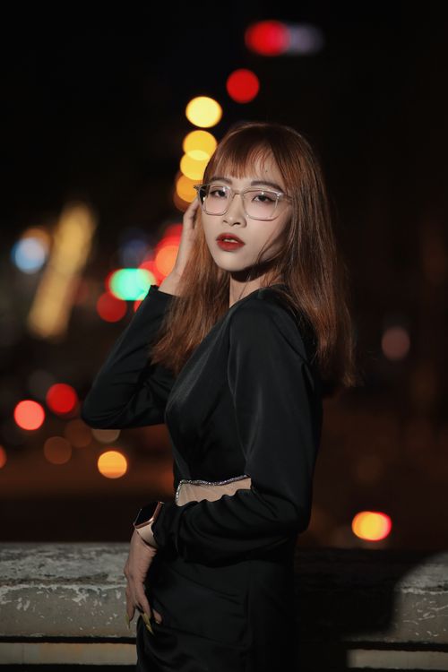 Free Young woman in a black dress with glasses poses stylishly against urban night lights bokeh. Stock Photo