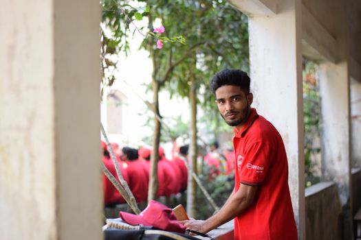 A young man in a red uniform smiles in an outdoor setting with greenery in the background.