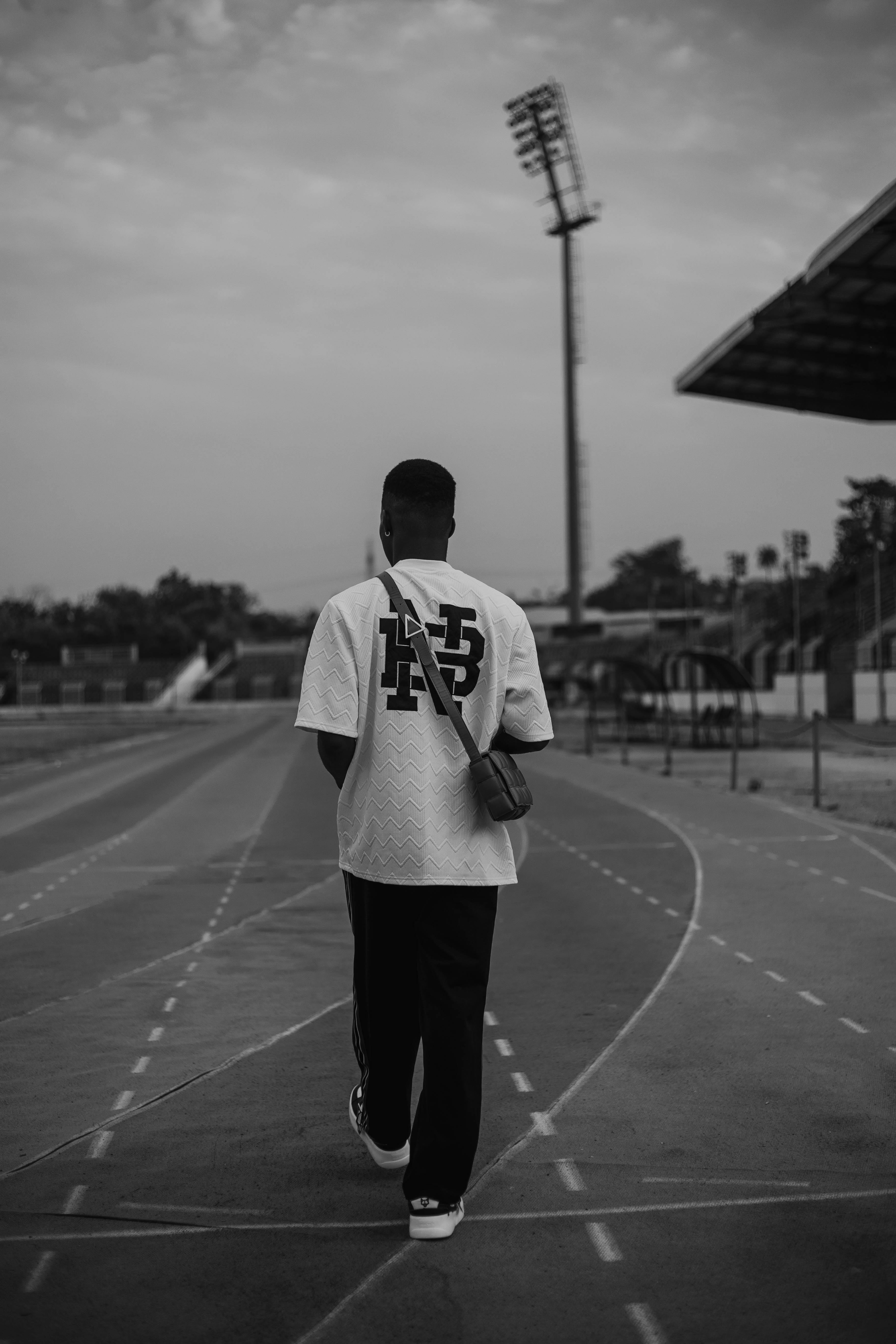 Grayscale photo of a man in streetwear walking on an urban track, showcasing modern fashion.