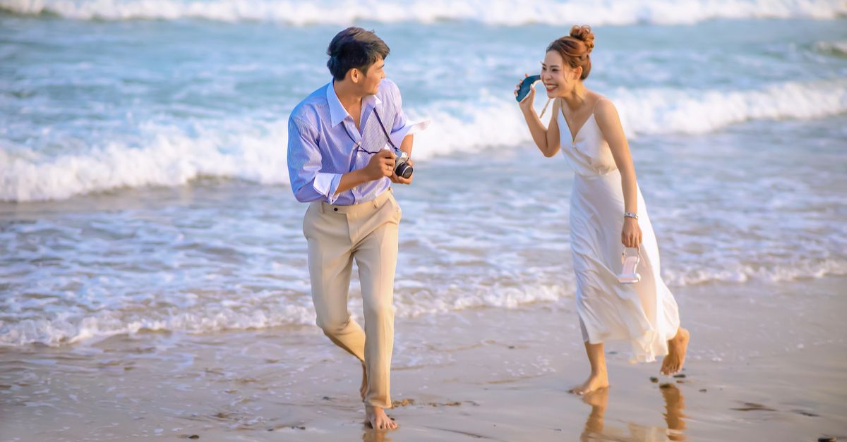 A joyful Asian couple shares a romantic moment while walking along a sunny beach.