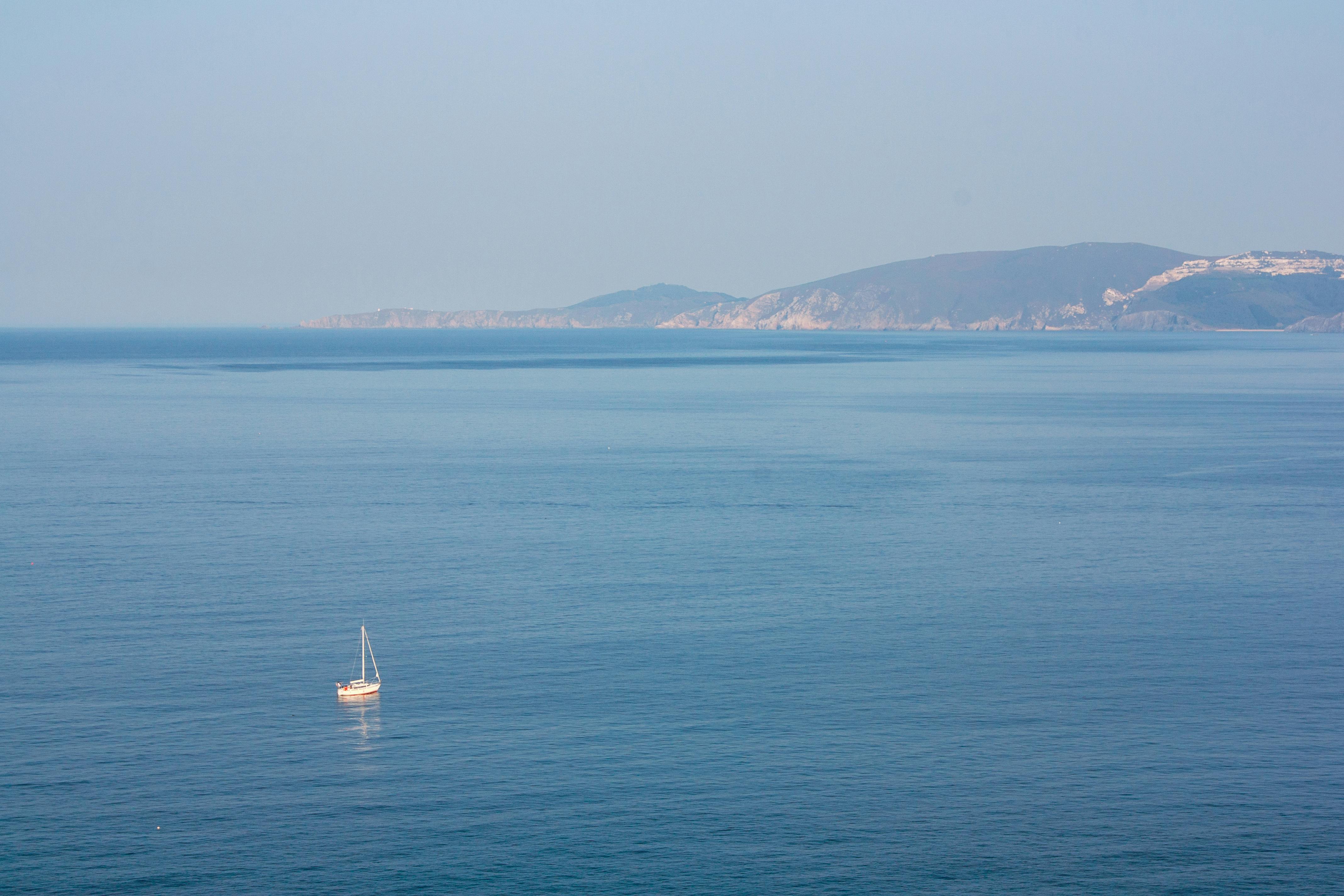 Serene aerial view of a lone sailboat on a vast blue ocean with distant cliffs under a clear sky.