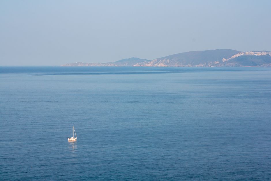 Serene aerial view of a lone sailboat on a vast blue ocean with distant cliffs under a clear sky.