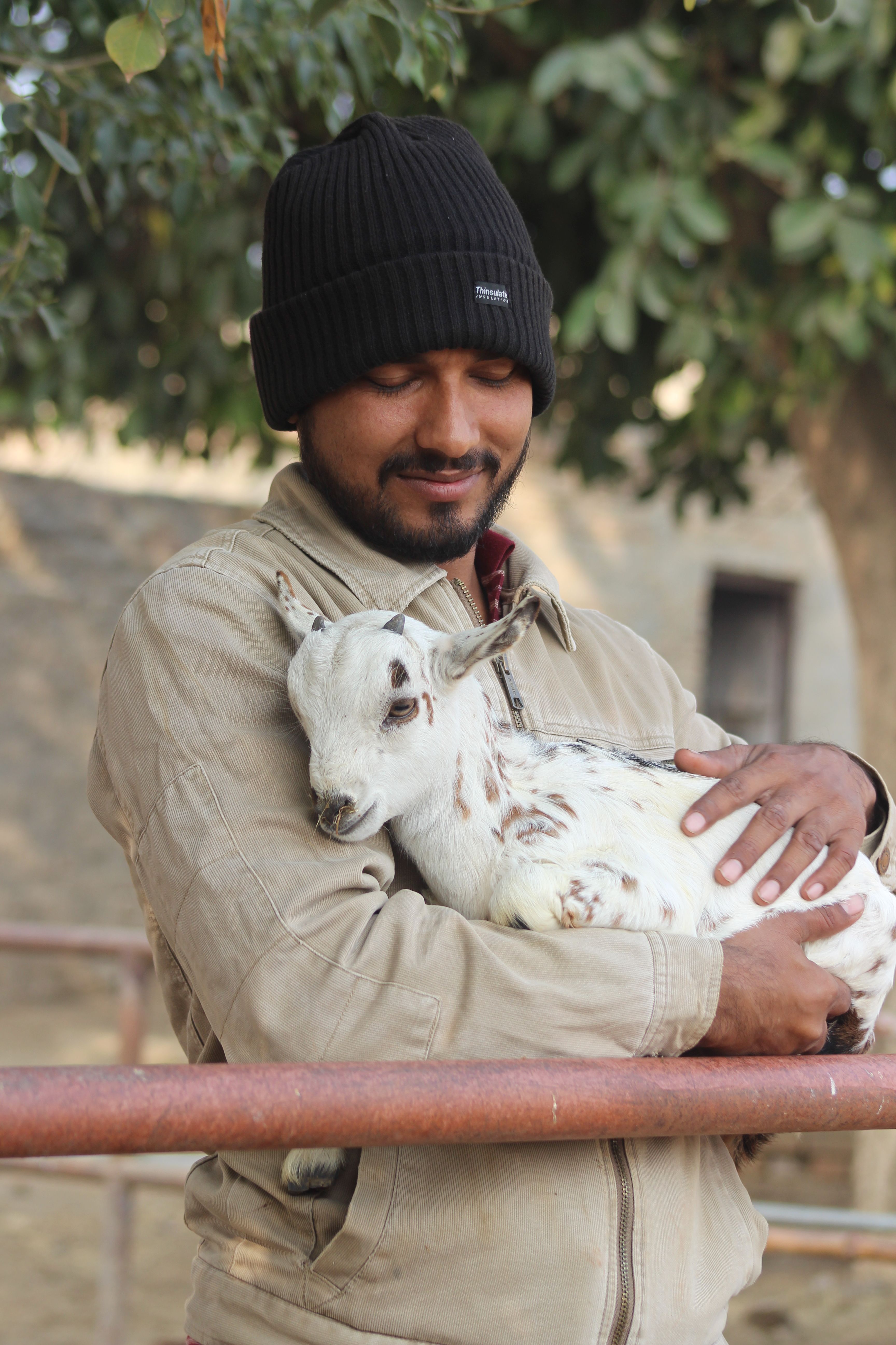 A man lovingly cradles a goat kid outdoors in a rural setting.