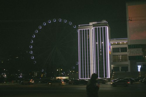 Free Cityscape featuring a Ferris wheel and illuminated building during nighttime, with cars and people. Stock Photo