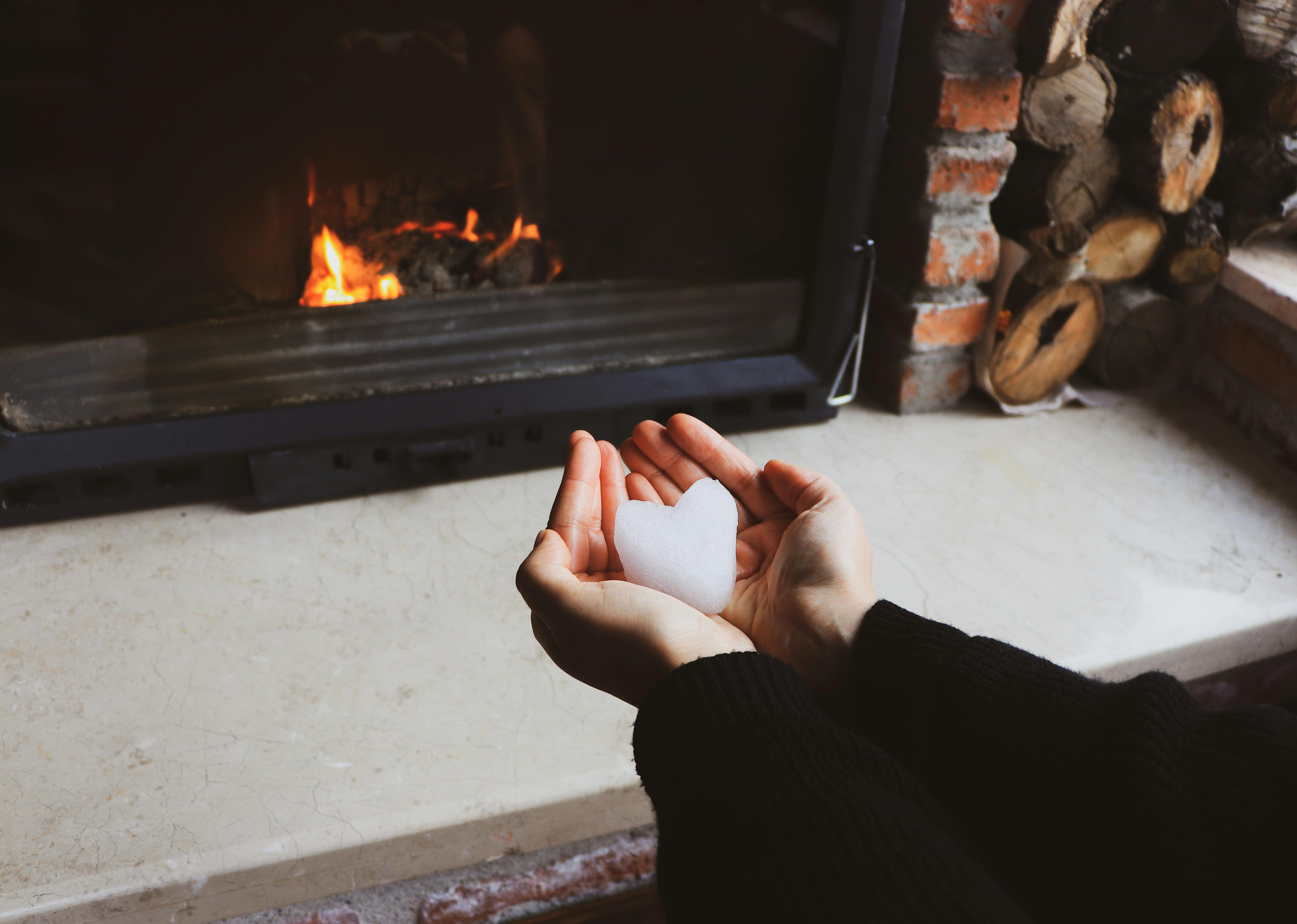Hands holding heart-shaped snow by a warm fireplace. Cozy winter indoor setting.