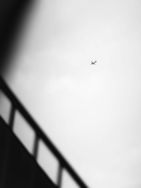 A minimalist black and white photo of an airplane flying overhead with a blurred wall in the foreground.