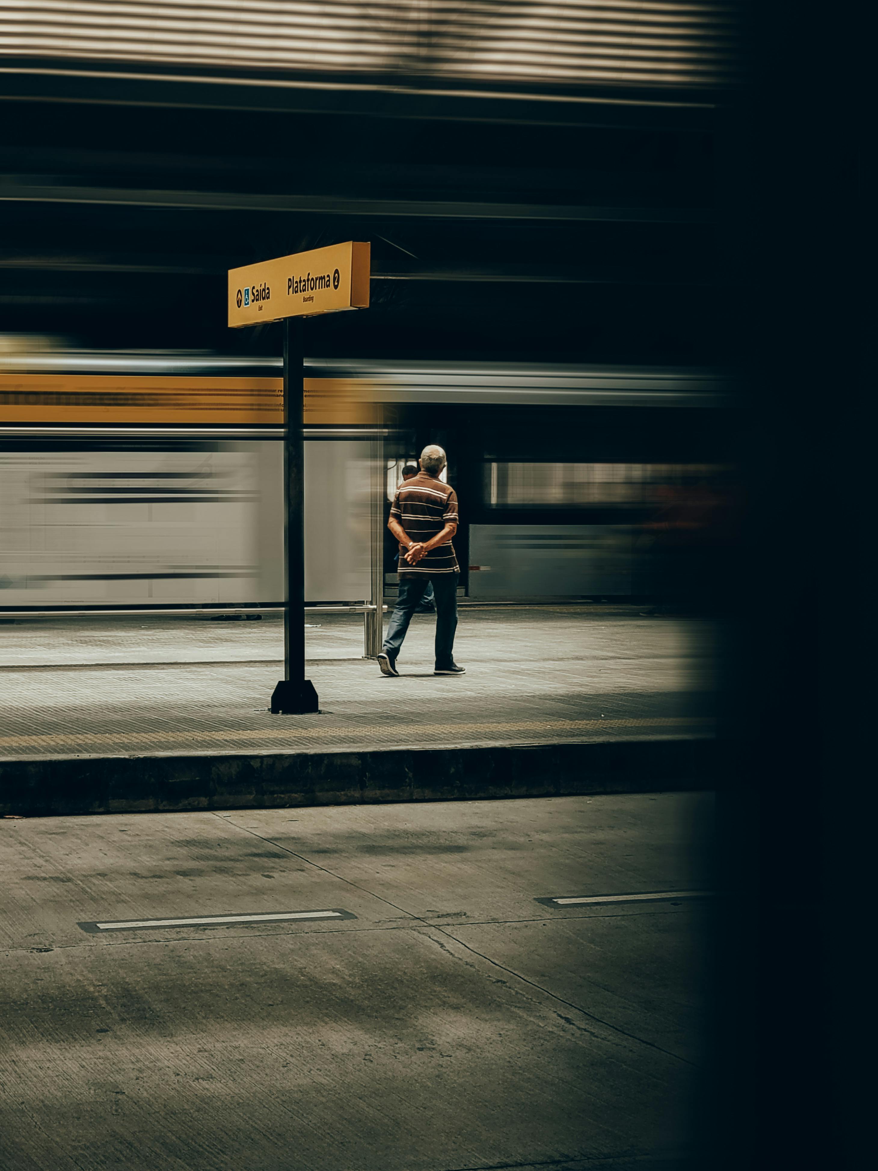 Man waits at urban railway platform with train in motion, creating a sense of blurred movement.