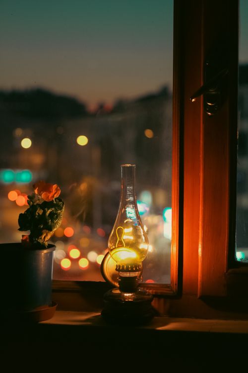 Free Warm evening glow through a window with a lantern and potted plant, capturing city bokeh lights. Stock Photo