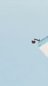 A low-angle view of a security camera on a blue and white building corner against a blue sky.