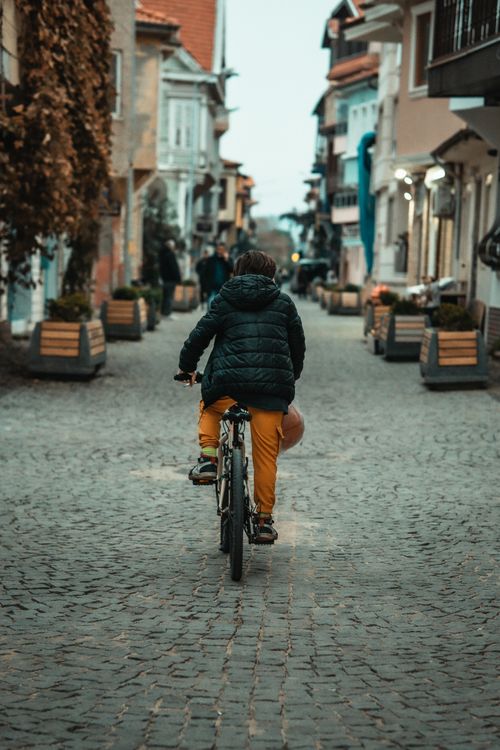 Free A young boy wearing a puffer jacket rides a bicycle down a cobblestone street, creating a charming urban scene. Stock Photo
