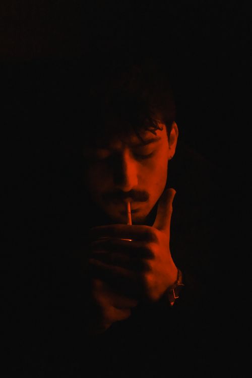 Free A contemplative young man lights a cigarette amidst dim red lighting, creating a moody atmosphere. Stock Photo