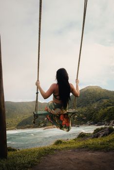 A woman with long hair sits on a swing, enjoying a serene view of the coastline and lush hills.
