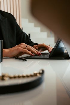 Close-up of a woman typing on a laptop in an office setting, showcasing hands with red nail polish.