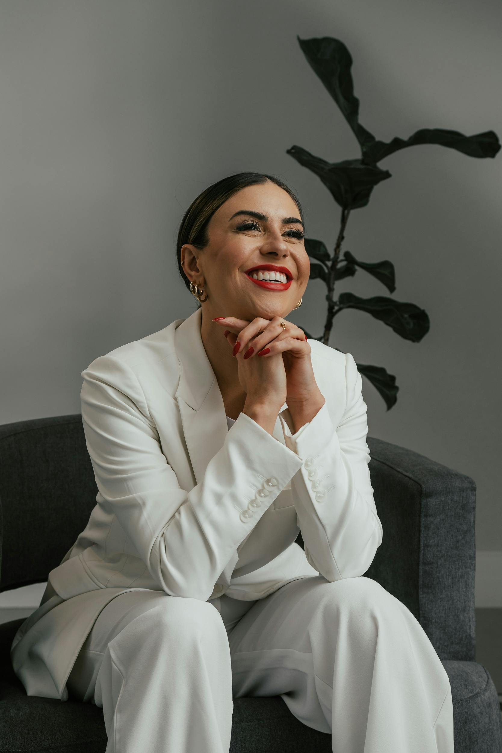 A confident woman in a stylish white suit smiles while sitting on a grey couch, exuding elegance and joy.