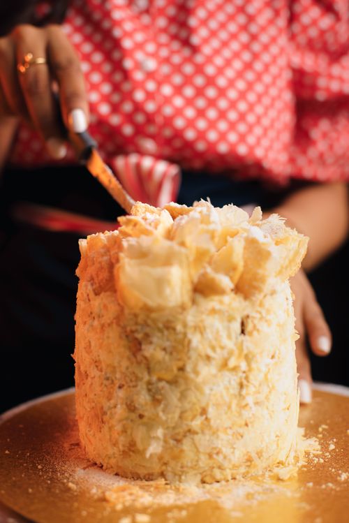 Free A person in a red dress cuts a tall Napoleon cake on a golden plate indoors. Stock Photo