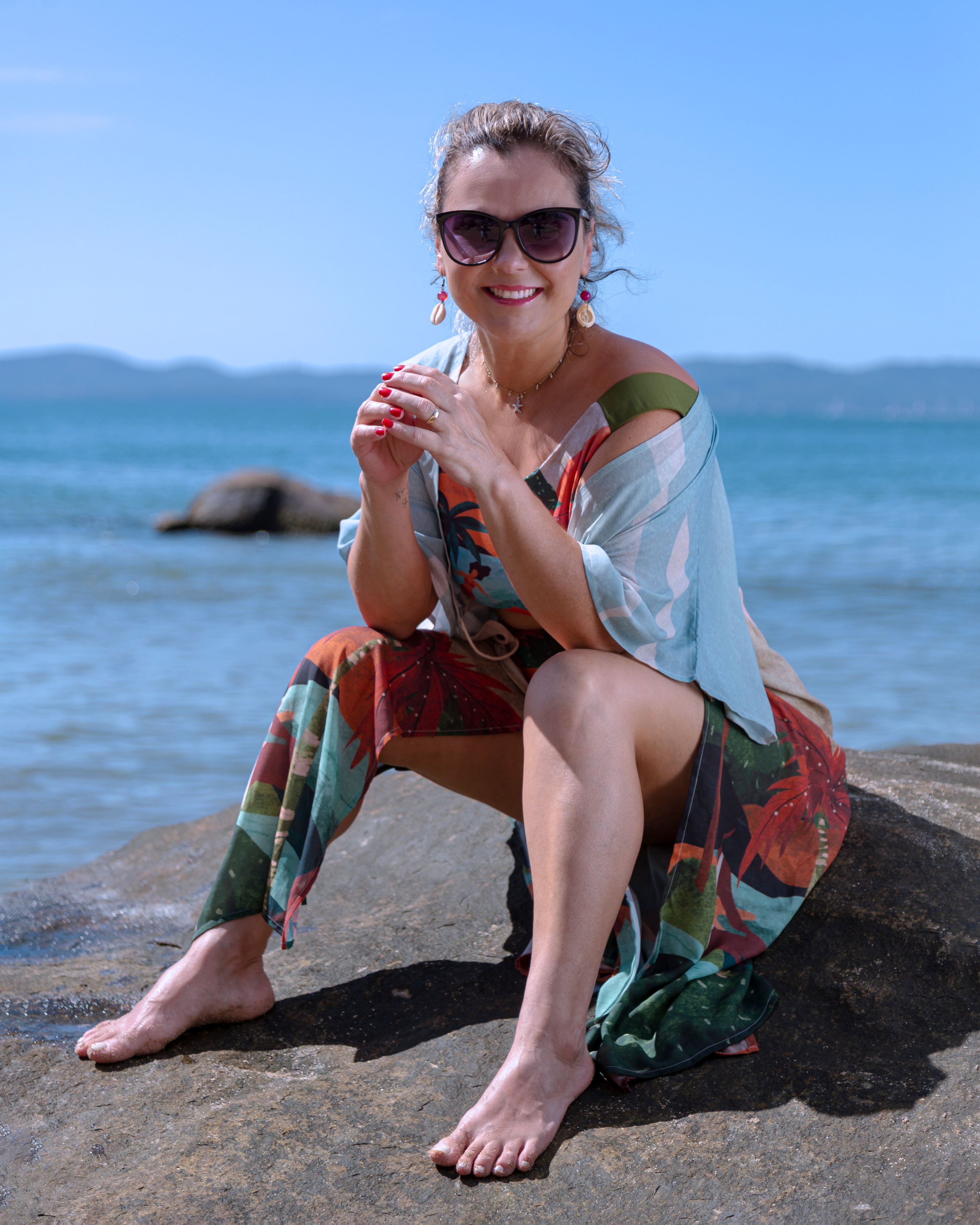 Mulher sorridente em um vestido colorido sentada em uma costa rochosa, aproveitando um dia ensolarado à beira do mar.