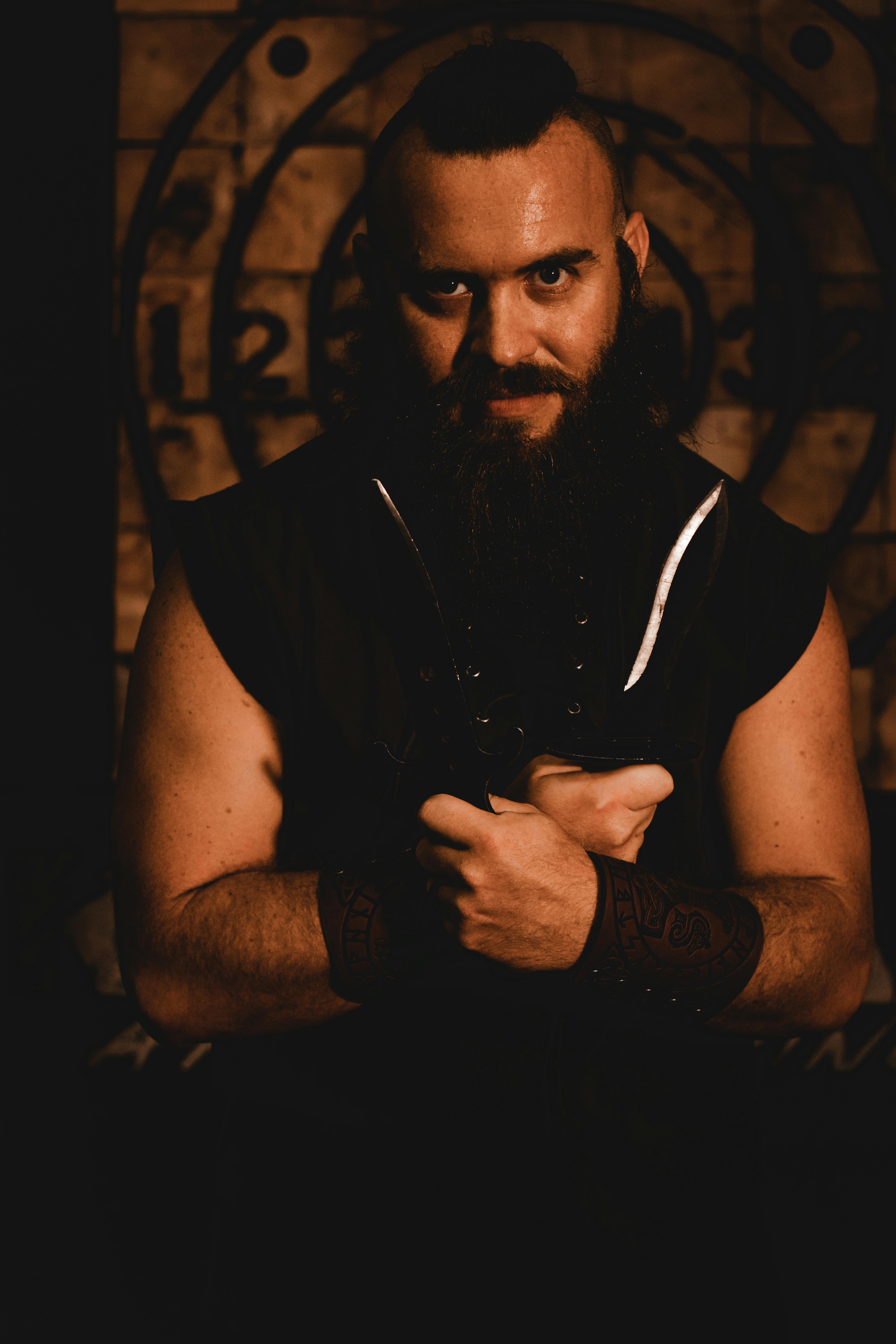 A muscular man with a beard holds knives in a dramatic, dimly lit portrait setting.
