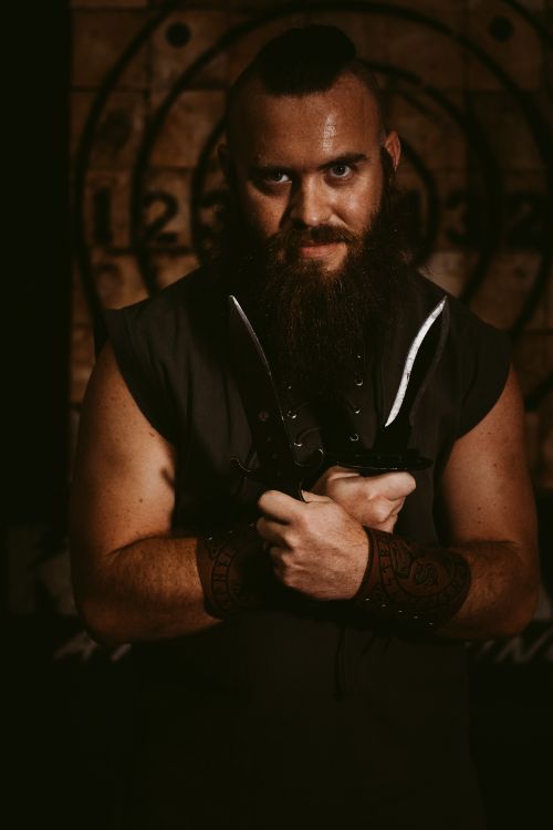 Free Bearded man with knives posing in a dimly lit room, exuding a mysterious aura. Stock Photo