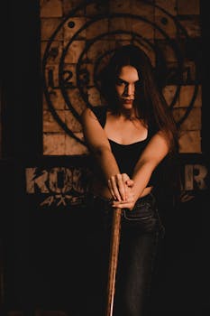 Portrait of a woman with long hair holding an axe against a dartboard background in moody lighting.