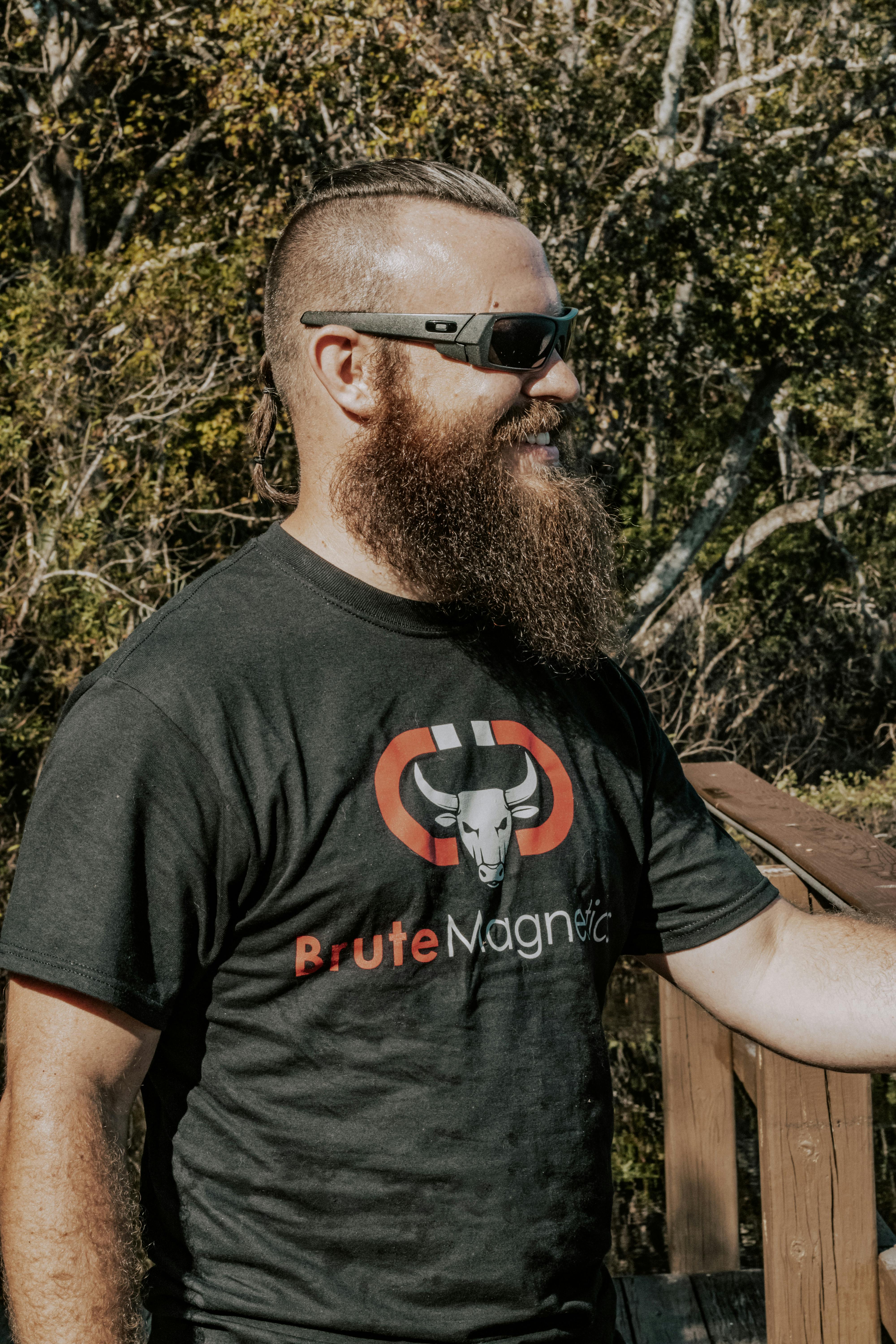 A fashionable man with a beard and sunglasses enjoys the outdoors in a casual t-shirt.