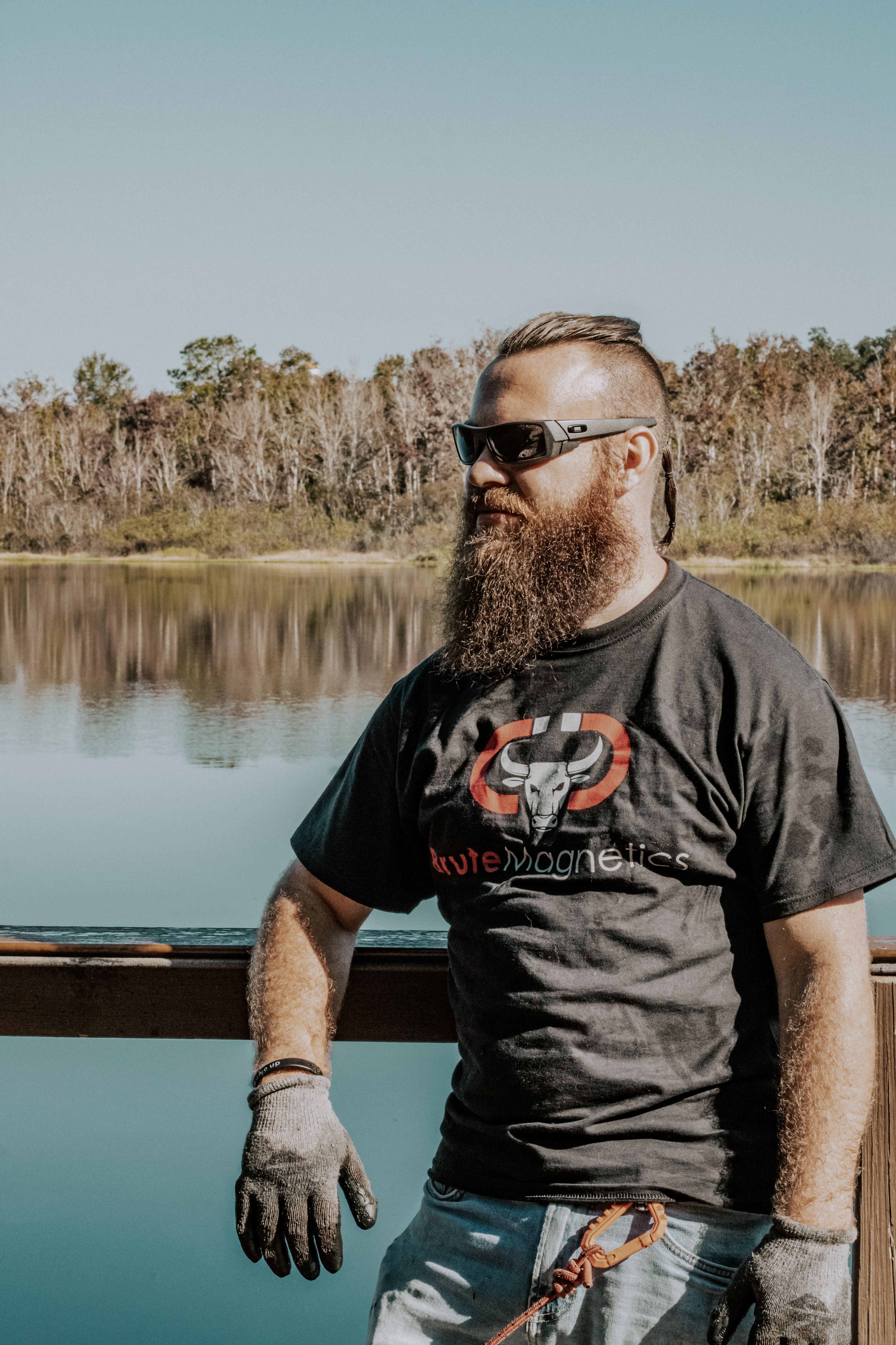 A bearded man wearing sunglasses poses by a tranquil lakeshore on a sunny day.
