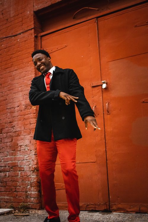 Free Fashionable man in vibrant red trousers and black coat poses against a rustic orange door in an urban alley. Stock Photo