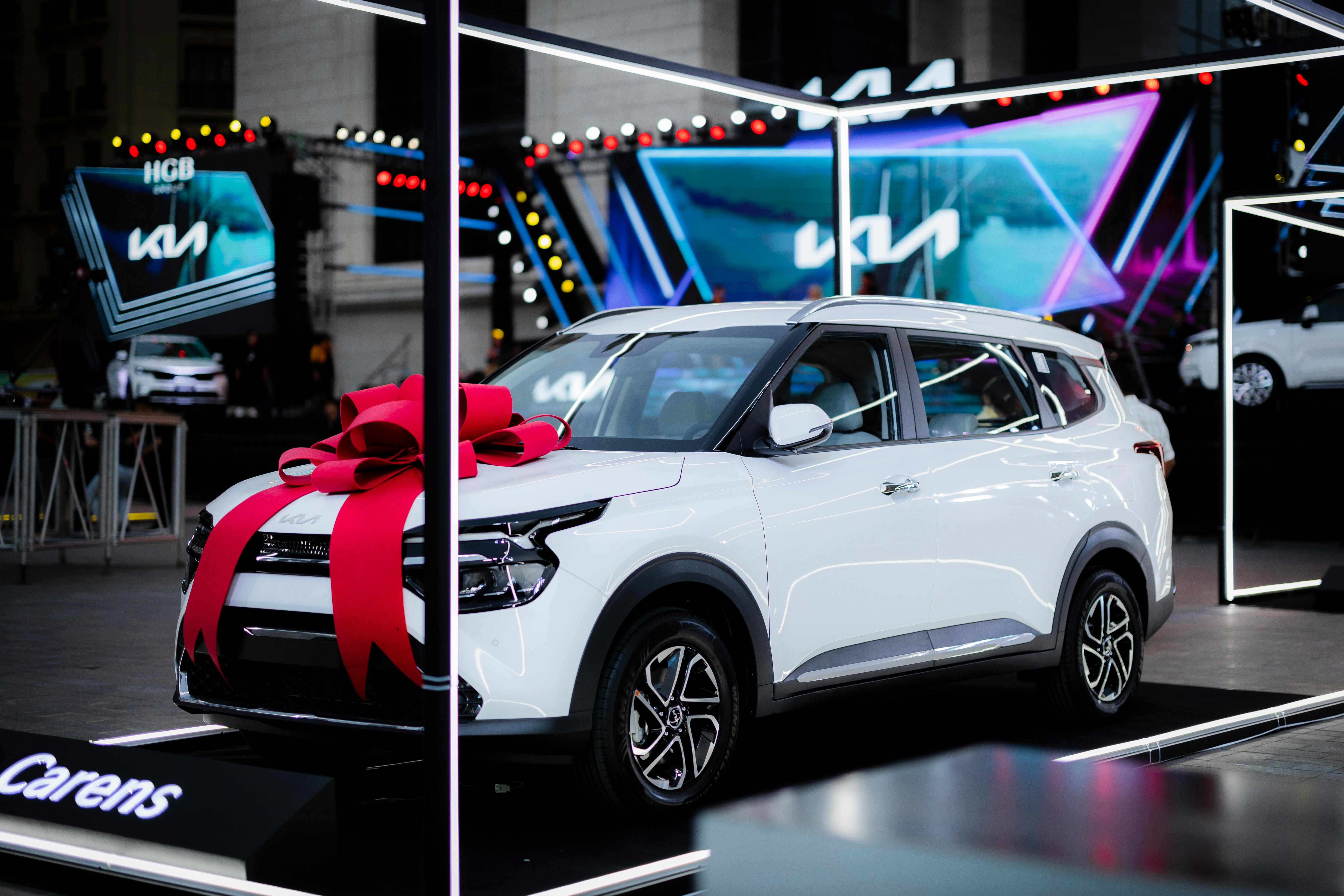 White car with a red bow on display at an automotive exhibition under bright lights.