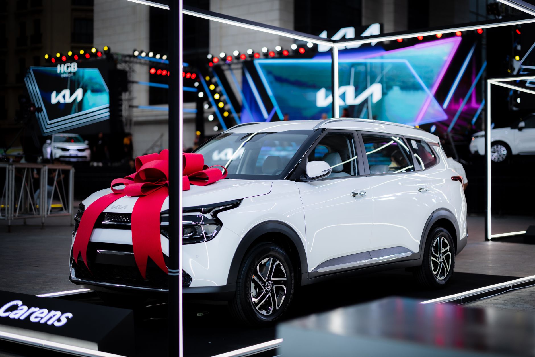 White car with a red bow on display at an automotive exhibition under bright lights.