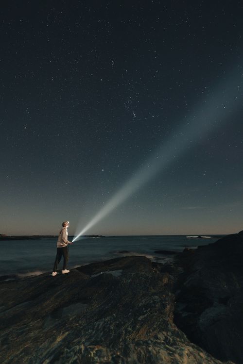 Free Person exploring rocky shoreline at night in Newport, RI, under starry sky with flashlight. Stock Photo