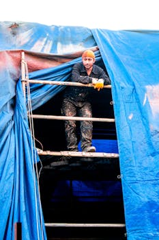 A construction worker stands on scaffolding wrapped in a blue tarp, wearing protective gear.