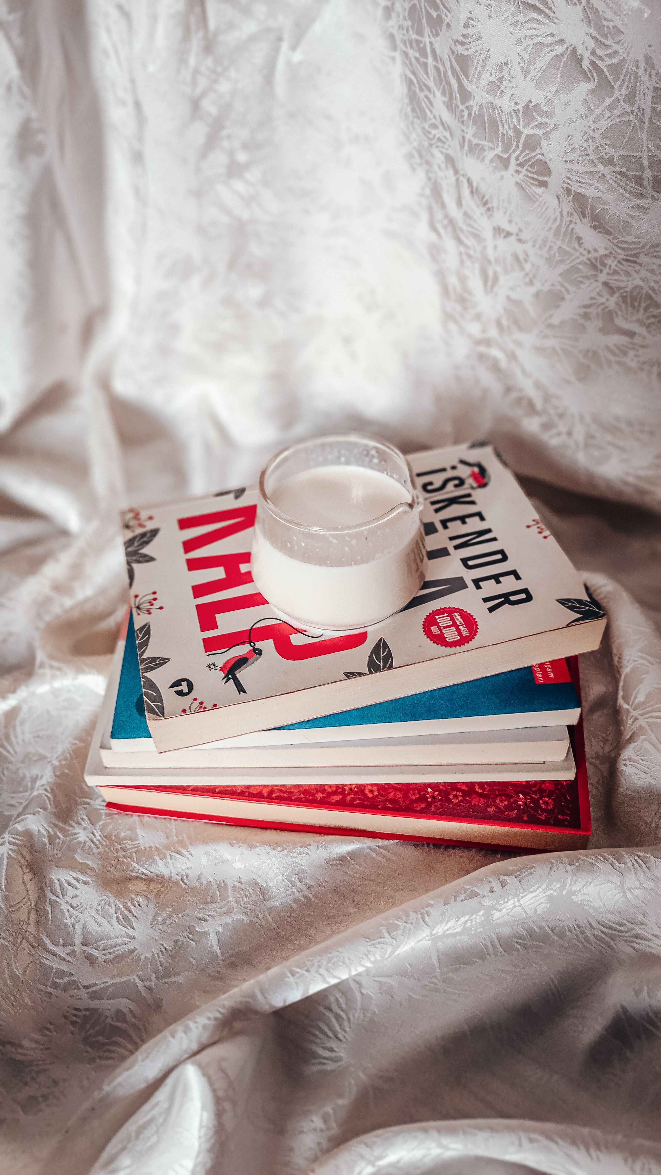 A stack of colorful books with a glass of milk on a draped fabric background.