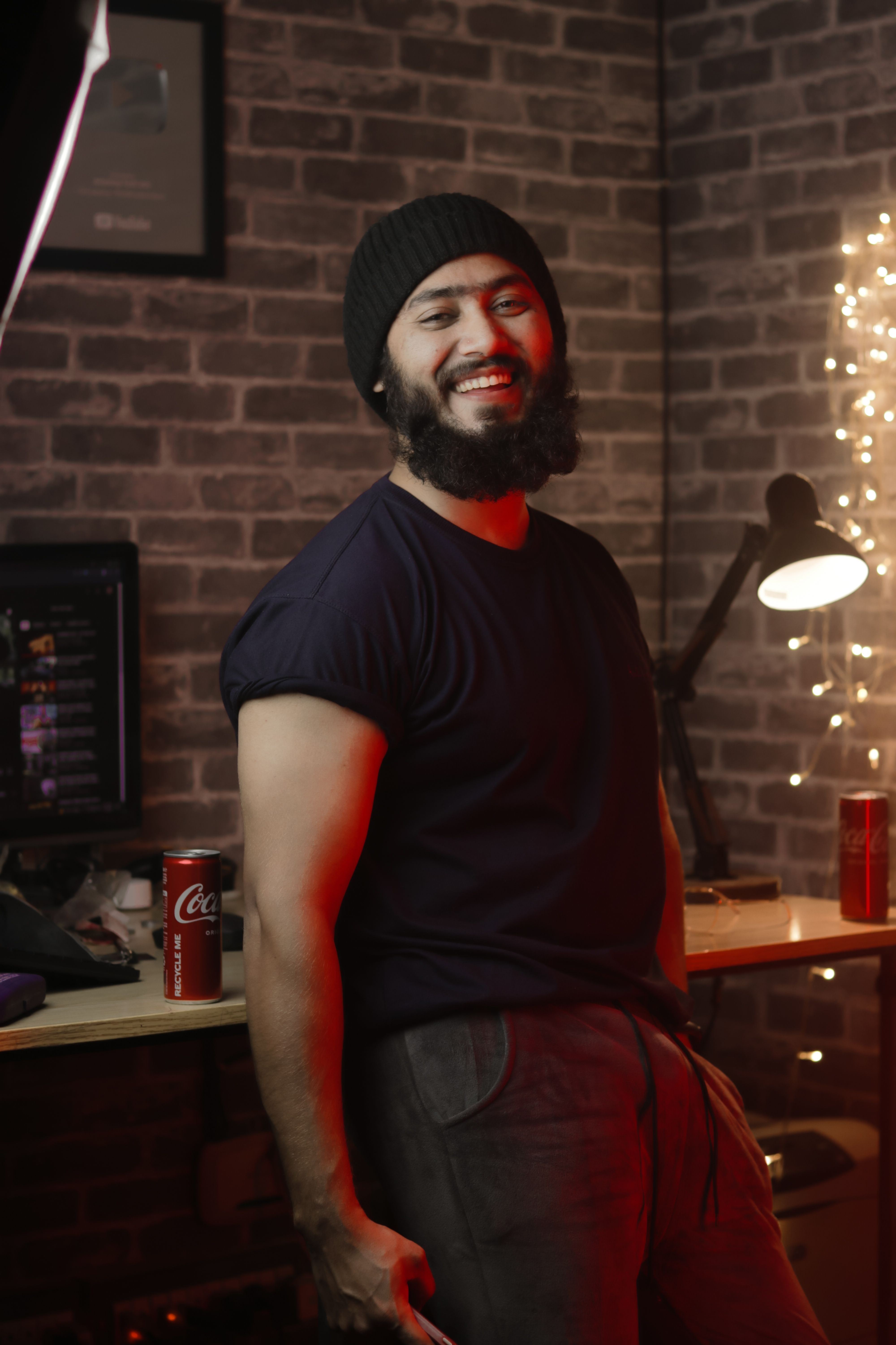 A bearded man in a cozy room smiles while leaning against a desk with soft lighting.